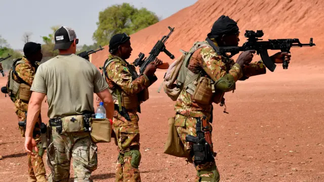 US military instructor passes by Malian soldiers holding weapons in camouflage gear, taken in 2018.