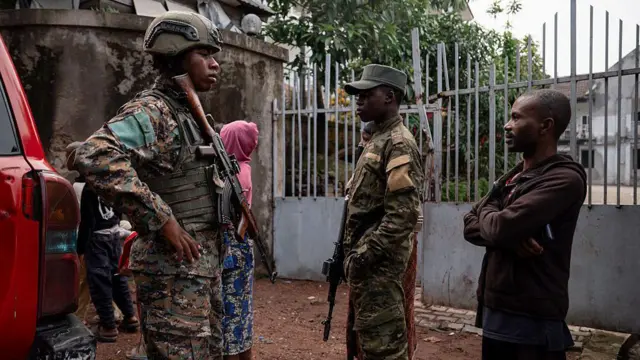 Two military personnel in uniform with rifles stand beside a civilian in a brown hoodie and pants, with a woman in a pink hoodie in the background